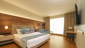 A hotel room with a large bed, a desk, a chair, a TV mounted on the wall, and a window with curtains. - Indus Biznotel (New Delhi)