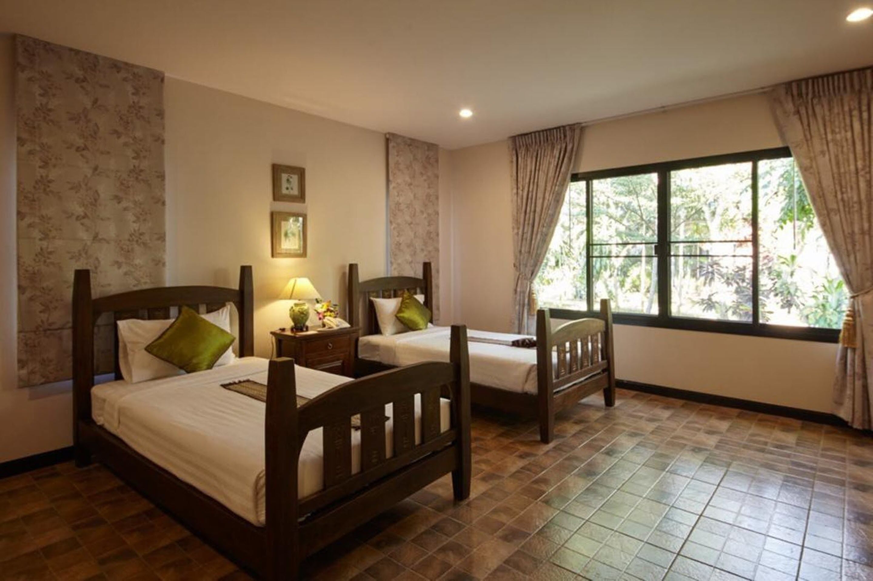 Una habitación con dos camas de madera, una mesita de noche con lámpara, una silla y un ventanal con cortinas.