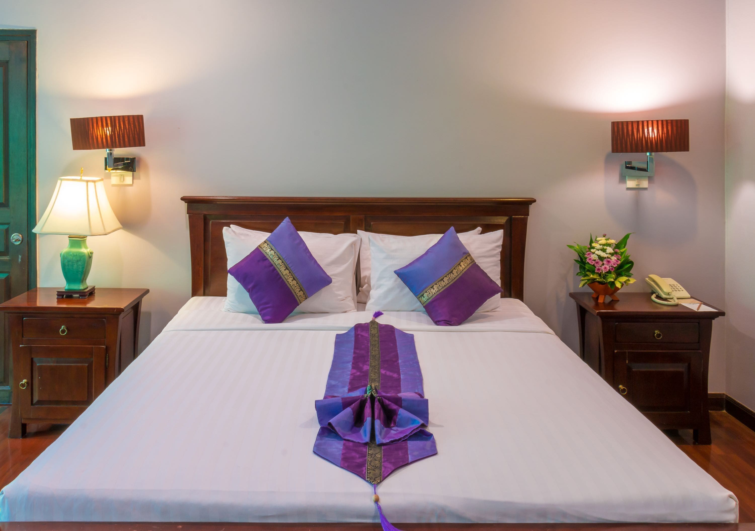 Un lit bien fait, agrémenté d’oreillers violets et blancs, d’un ruban décoratif et d’une tête de lit en bois. On y trouve deux tables de chevet avec des lampes, et une petite plante sur l’une d’elles.