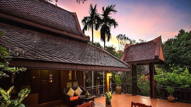 A traditional building with a tiled roof, a covered patio with wooden furniture, and a view of palm trees. - Phuwanalee Resort (Pak Chong)