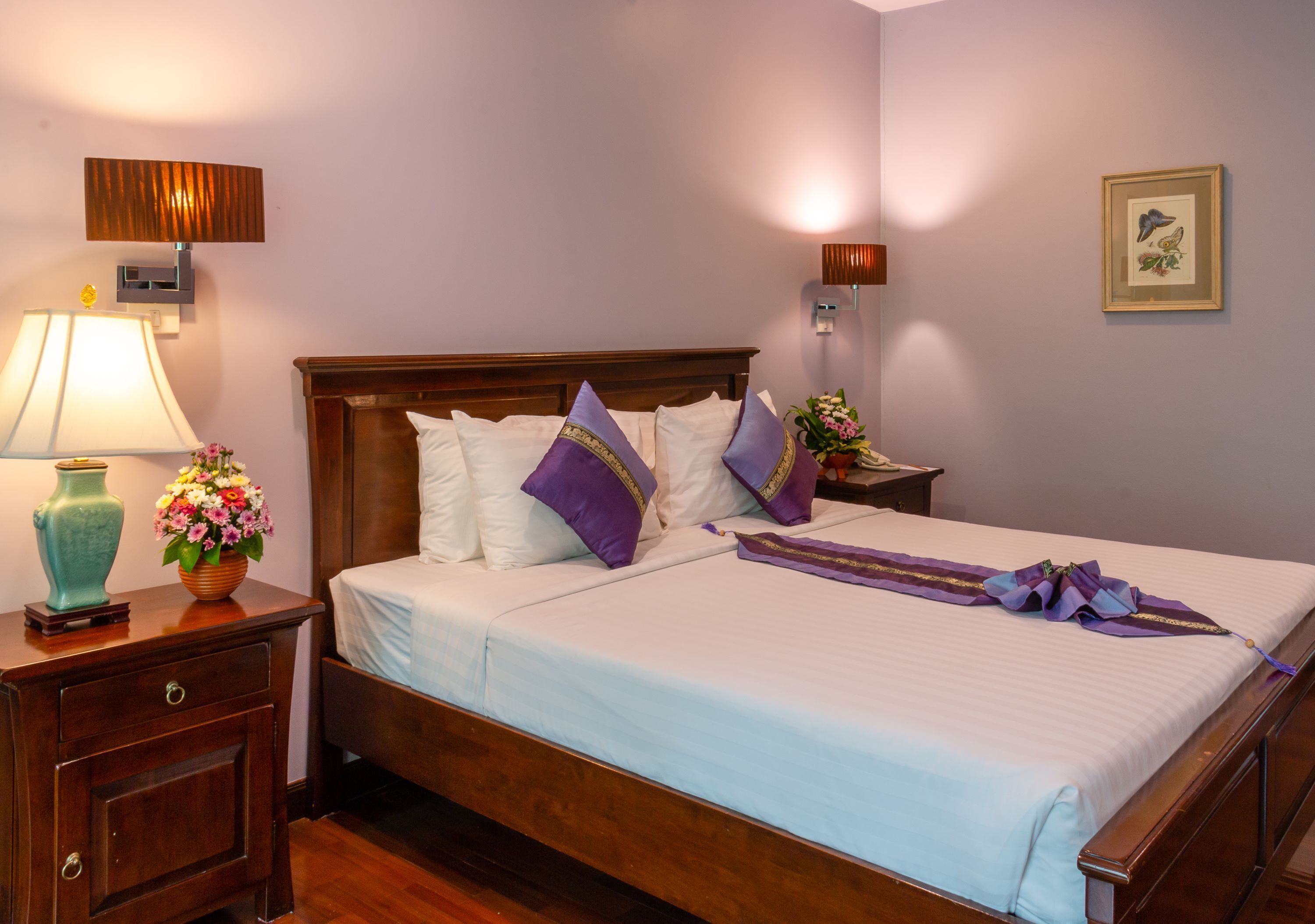 A neatly made bed with white and purple bedding, a wooden headboard, and a bedside table with a lamp and vase of flowers.