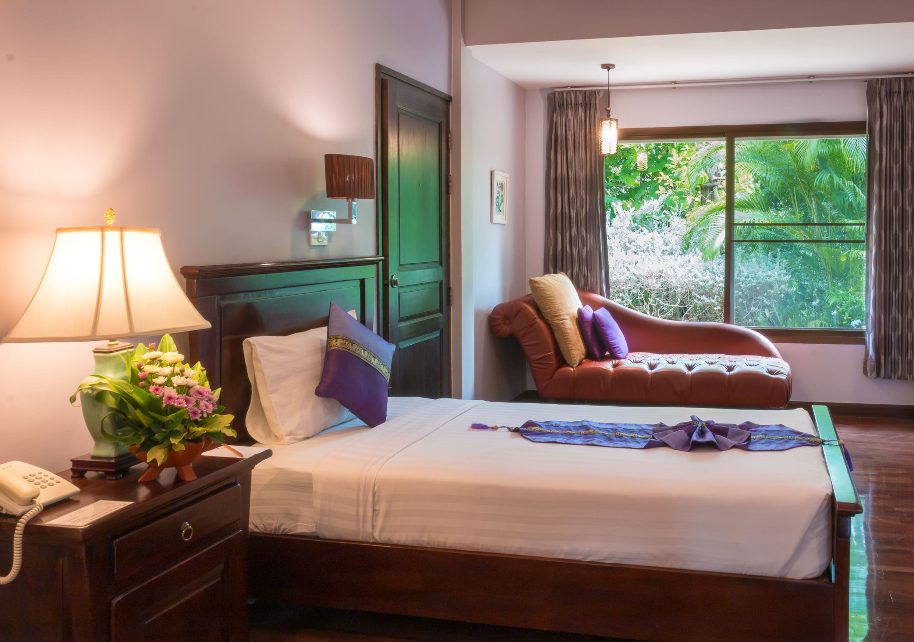A hotel room with a bed, a desk with a lamp and flowers, a chair, a window with curtains, and a view of greenery outside.