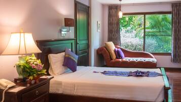 A hotel room with a bed, a desk with a lamp and flowers, a chair, a window with curtains, and a view of greenery outside.