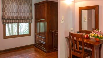 A room with wooden flooring, a large window with a patterned blind, a wooden cabinet, a desk with a mirror, and a vase with flowers.