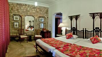 A hotel room with a brick wall, two beds with red floral bedspreads, wooden headboards, a small round table with chairs, and framed pictures on the wall.