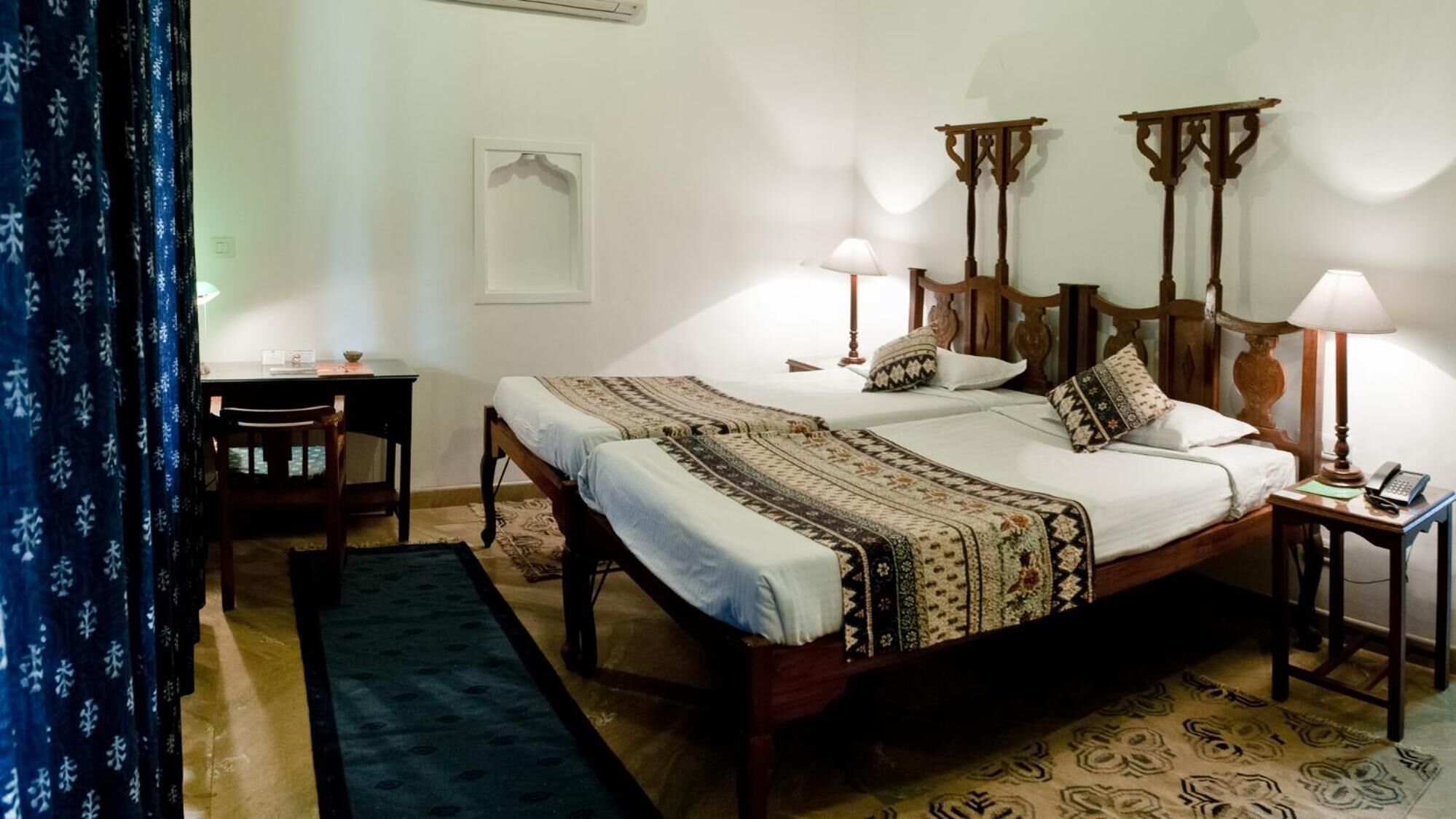 A double bed with a patterned bedspread, two bedside tables with lamps, a small desk with a chair, and a blue curtain.