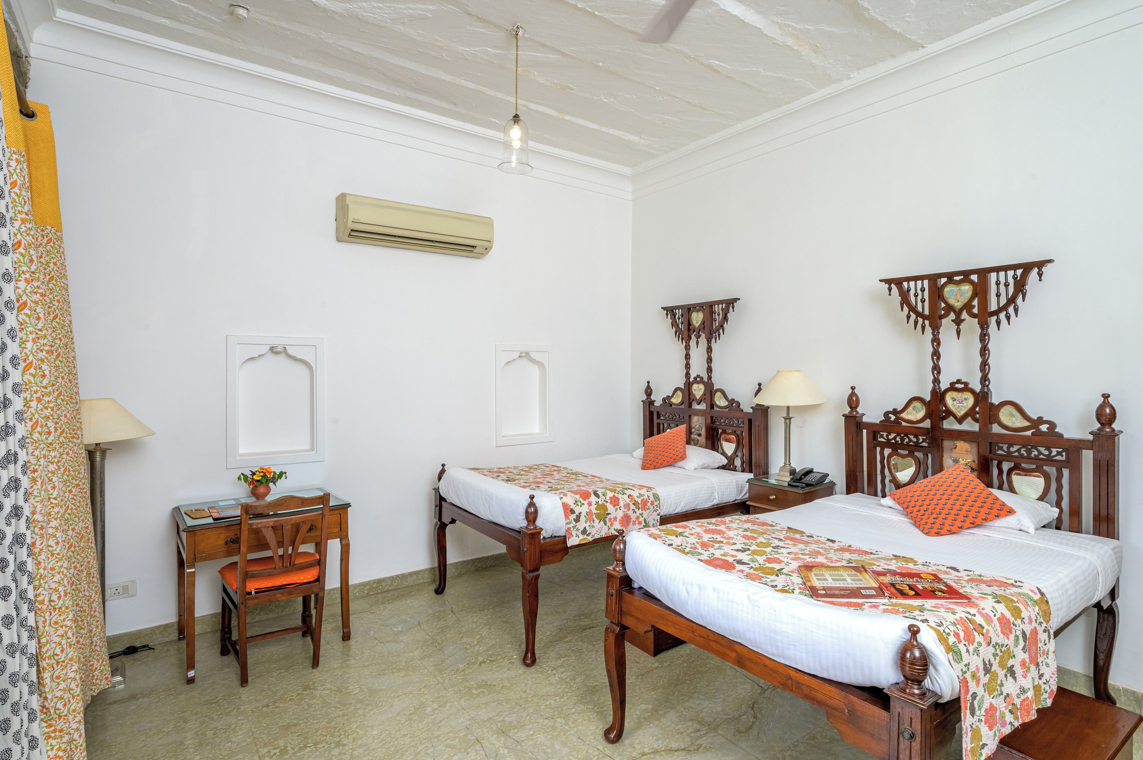 A room with two wooden beds, a desk, and a chair. There is an air conditioning unit on the wall and a hanging light fixture.