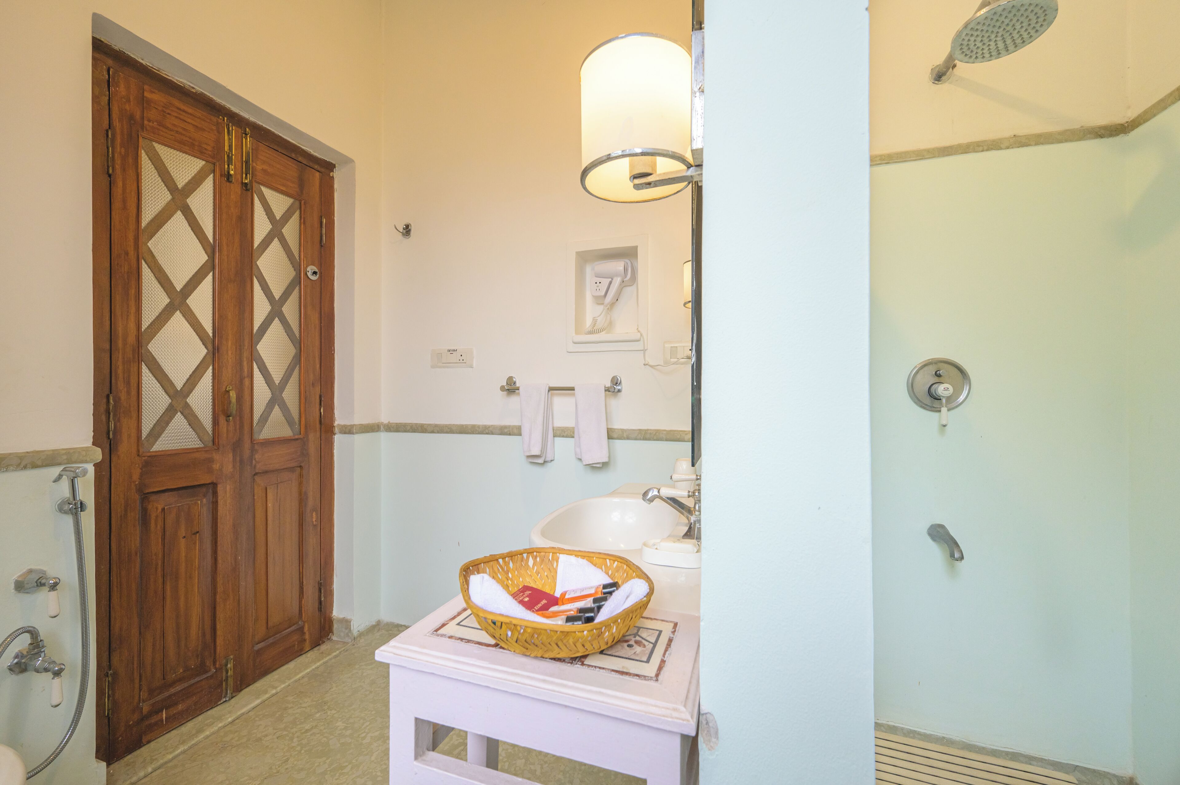 A bathroom with a wooden door, a white sink, a mirror, a shower, and a basket on the sink.