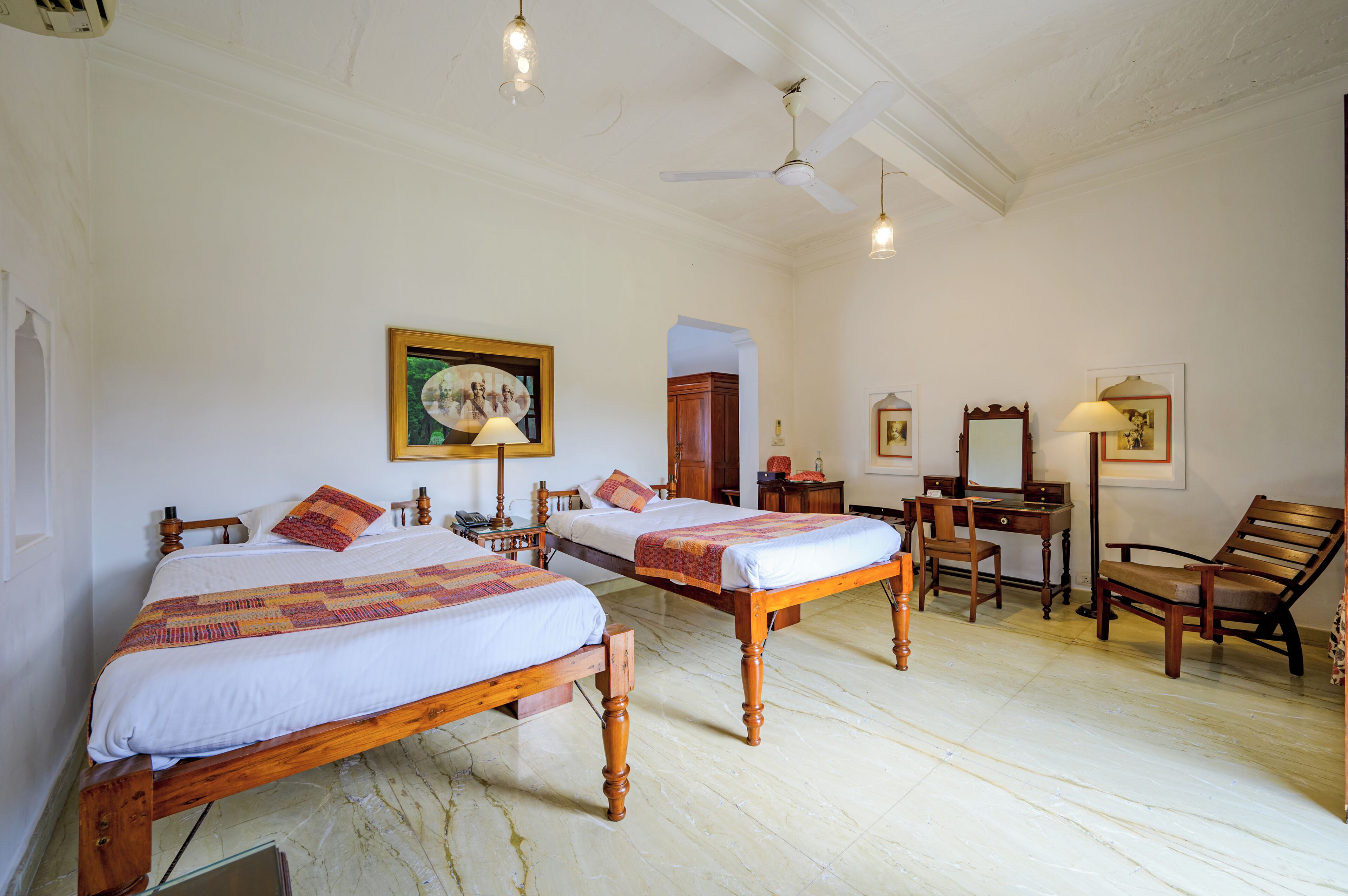 Une chambre avec deux lits en bois, un bureau, une chaise, un ventilateur de plafond et un tableau encadré au mur.