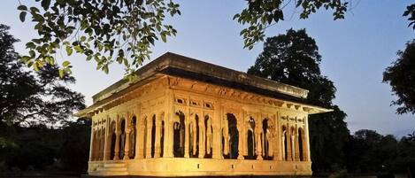 A well-lit, intricately carved stone structure with columns, surrounded by trees and a body of water.