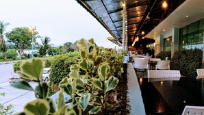 An outdoor seating area with white chairs and tables, surrounded by greenery and a view of the street. - The Viangtak Riverside Hotel (Tak)
