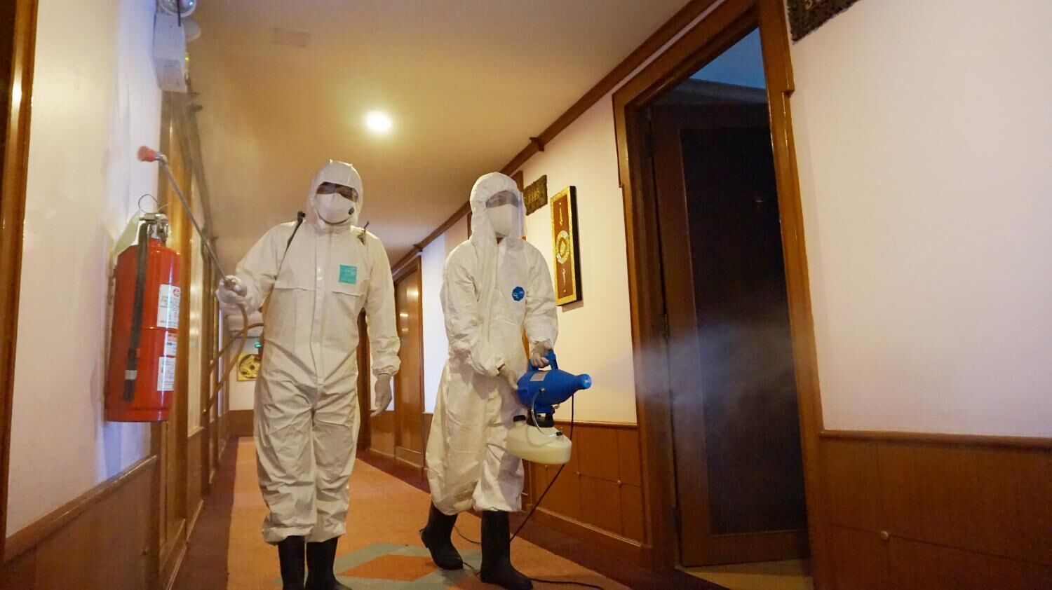 Two individuals in protective suits walking down a hallway with a fire extinguisher on the wall.
