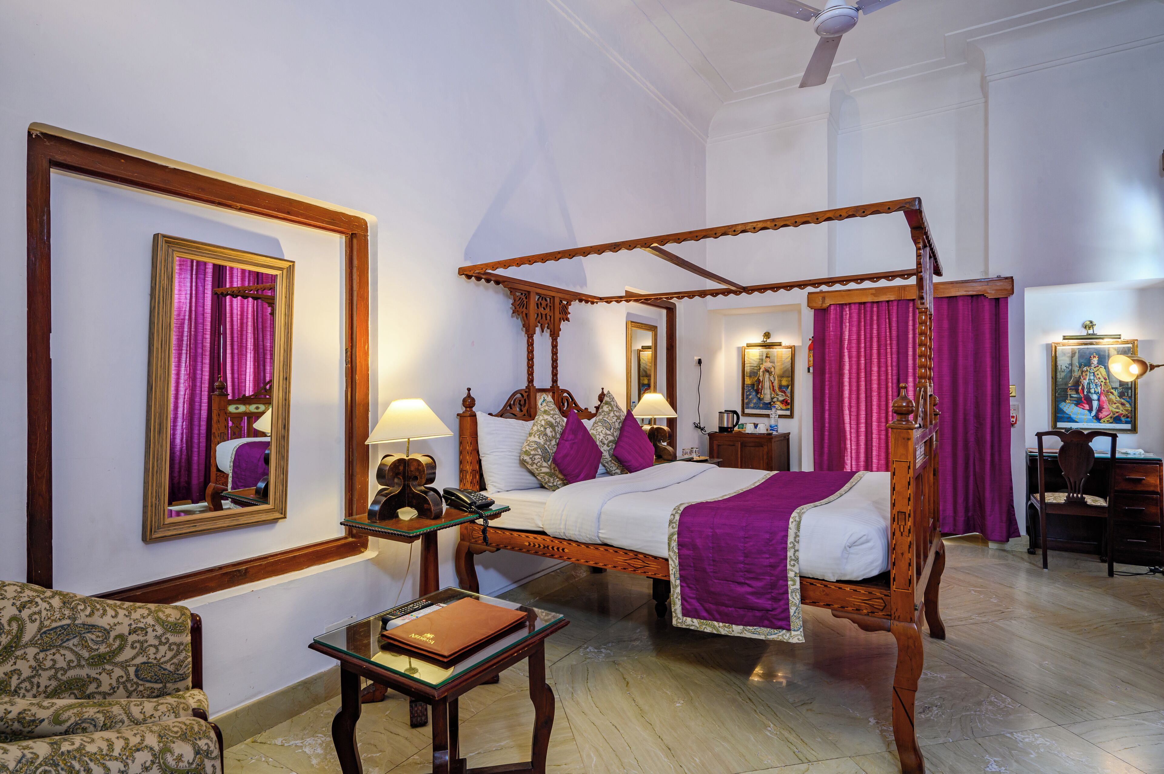 A four-poster bed with a wooden frame and purple bedding, a bedside table with a lamp, a chair, and a desk with a chair.