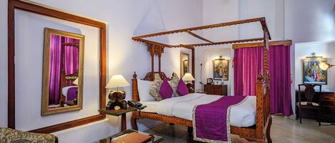 A four-poster bed with a wooden frame and purple bedding, a bedside table with a lamp, a chair, and a desk with a chair.