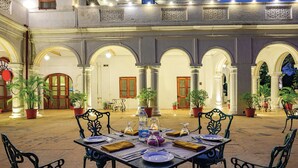 An outdoor dining area with tables set for two, surrounded by ornate metal chairs and potted plants. - Neemrana's Baradari Palace (Patiala)