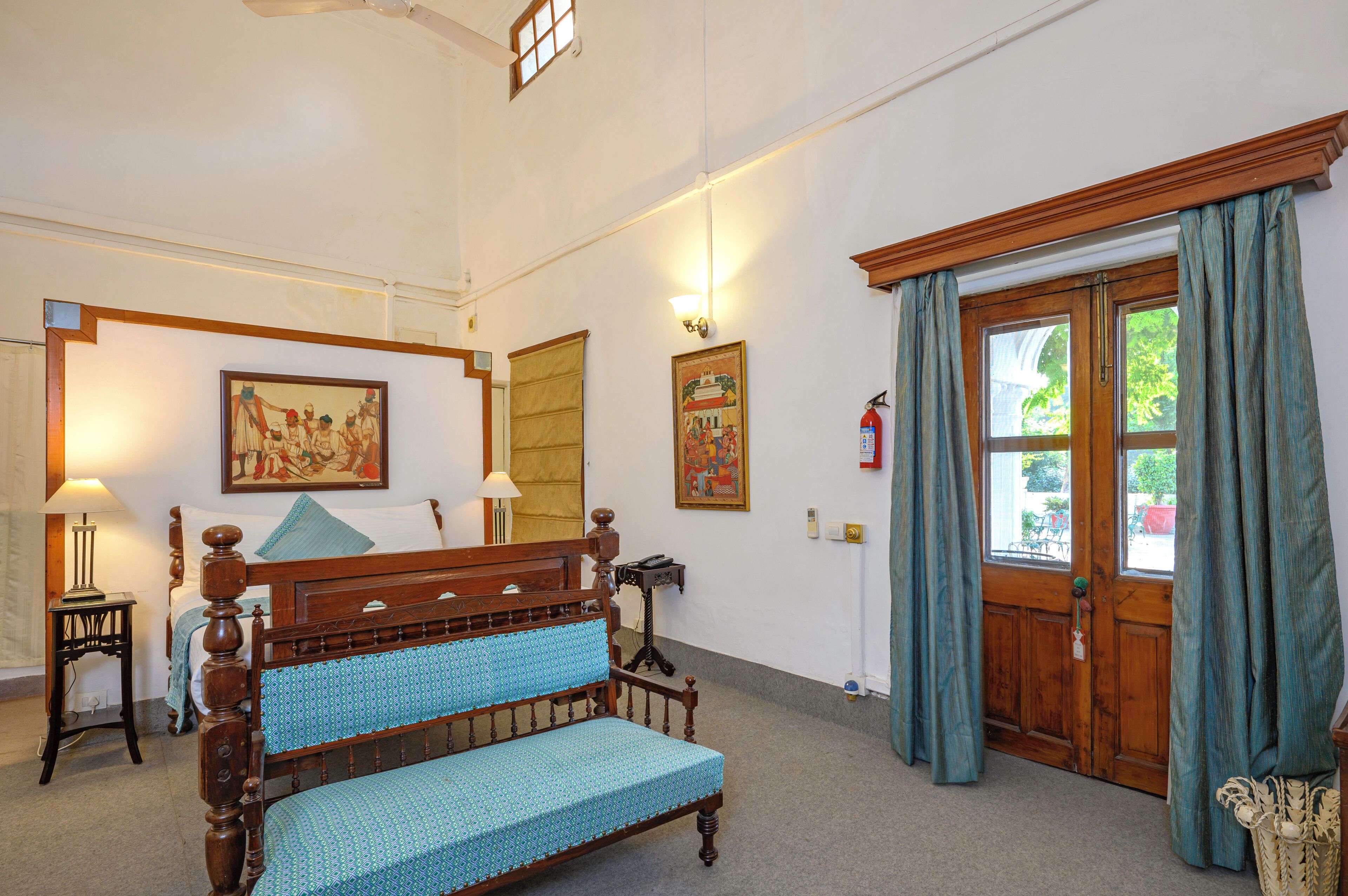 A room with a wooden bed, a wooden bench, a lamp, a framed picture, a door with curtains, and a window.