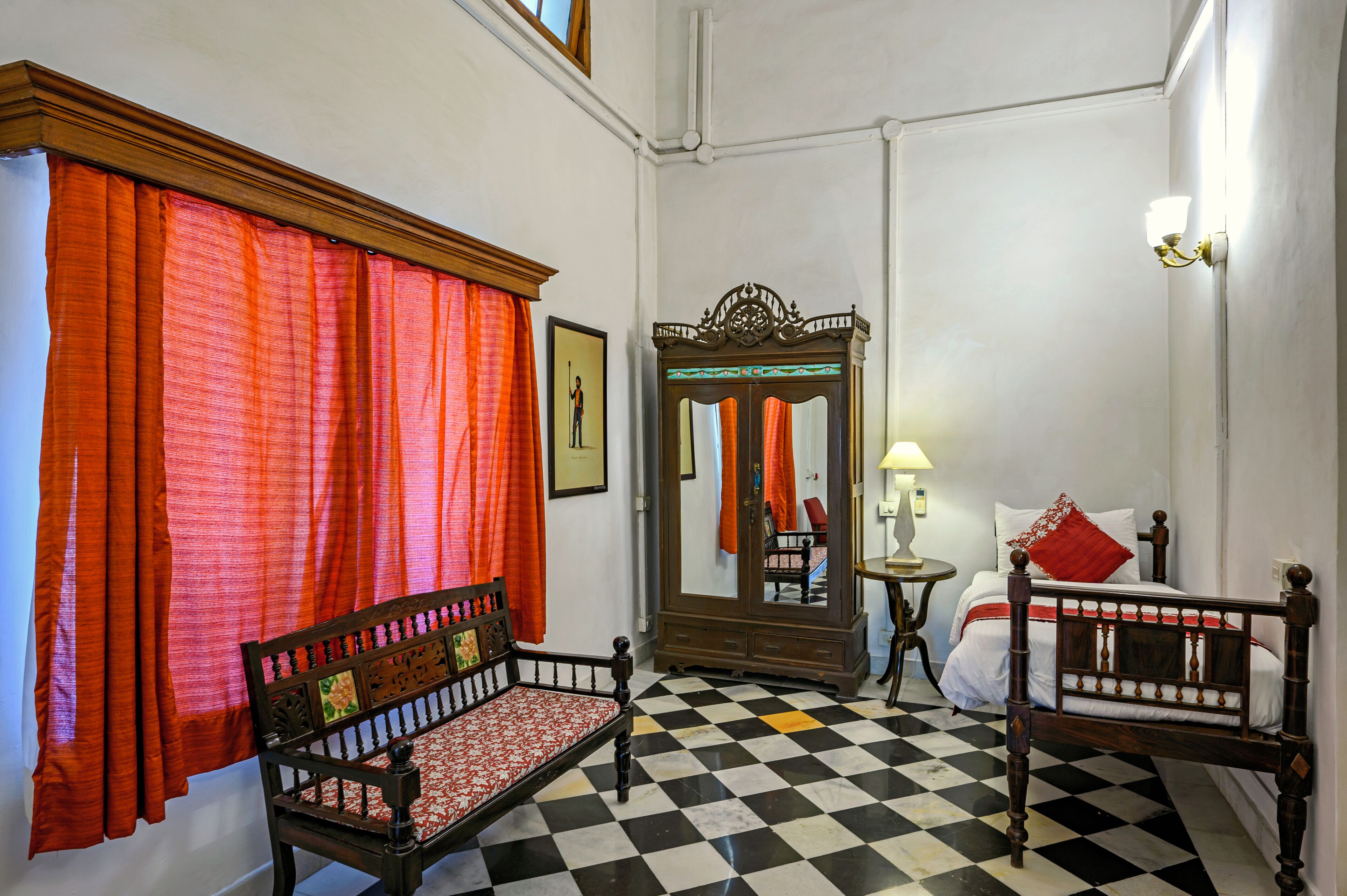 A room with a checkered floor, a wooden bed with white bedding, a wooden bench, a mirror, and red curtains.