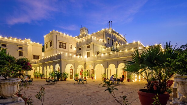 A courtyard with a building featuring arched walkways and string lights, surrounded by potted plants and outdoor seating. - Neemrana's Baradari Palace (Patiala)