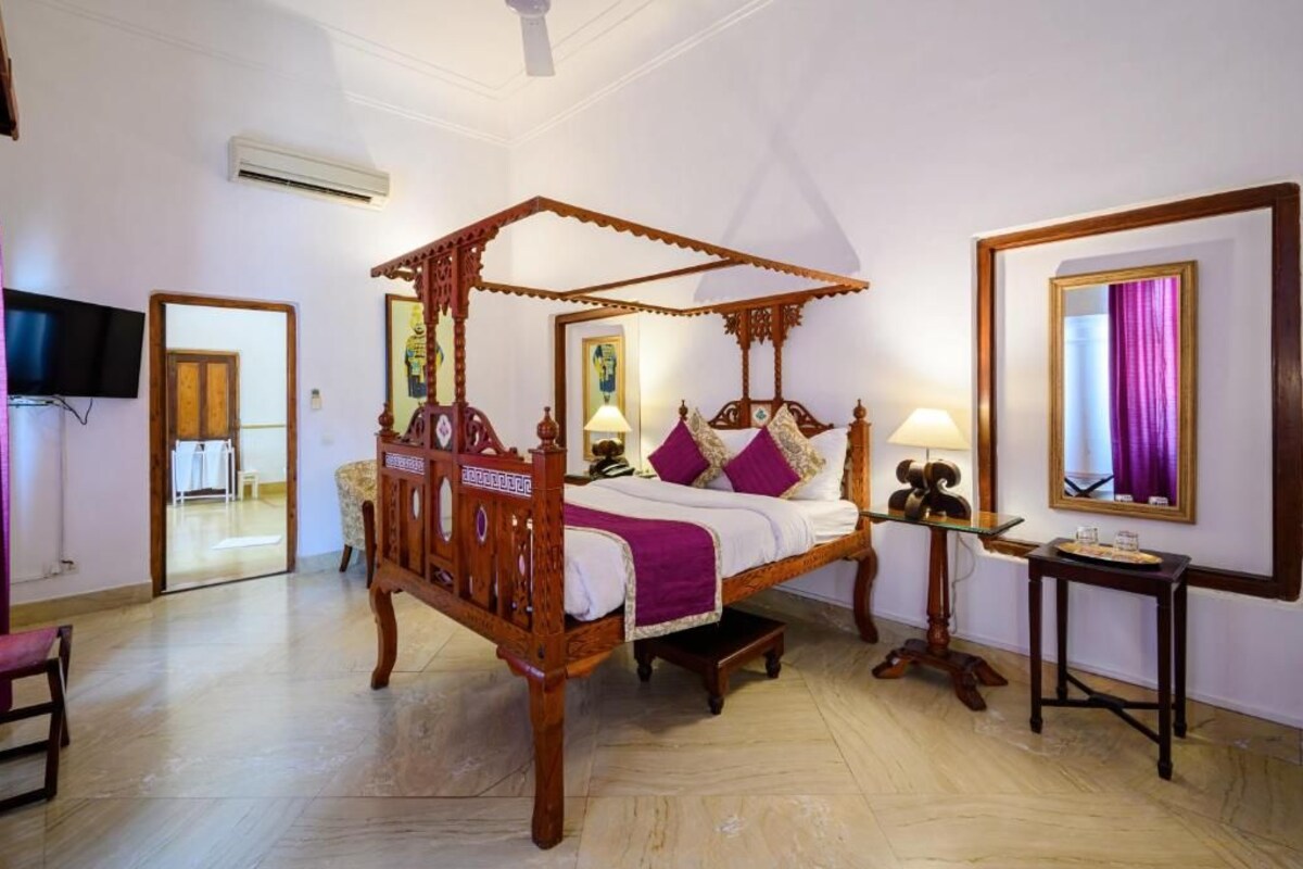 A four-poster bed with a wooden frame, a bedside table with a lamp, a mirror on the wall, and a television mounted on the wall.