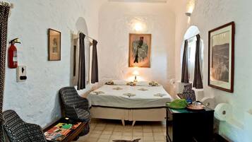 A small, cozy room with a bed, two chairs, a table with books, a lamp, and a fire extinguisher.