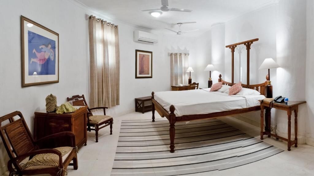 A hotel room with a large bed, a desk, two chairs, a mirror, and a ceiling fan.