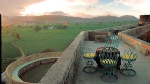 A terrace with a view of green fields and distant mountains, featuring a table and chairs. - Neemrana's - Hill Fort - Kesroli (Ramgarh)