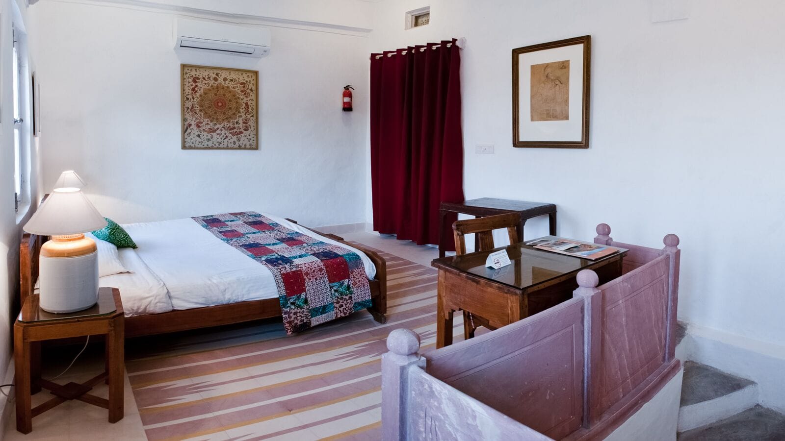 A hotel room with a bed, a bedside table with a lamp, a small table with a magazine, a chair, a red curtain, and a framed picture on the wall.