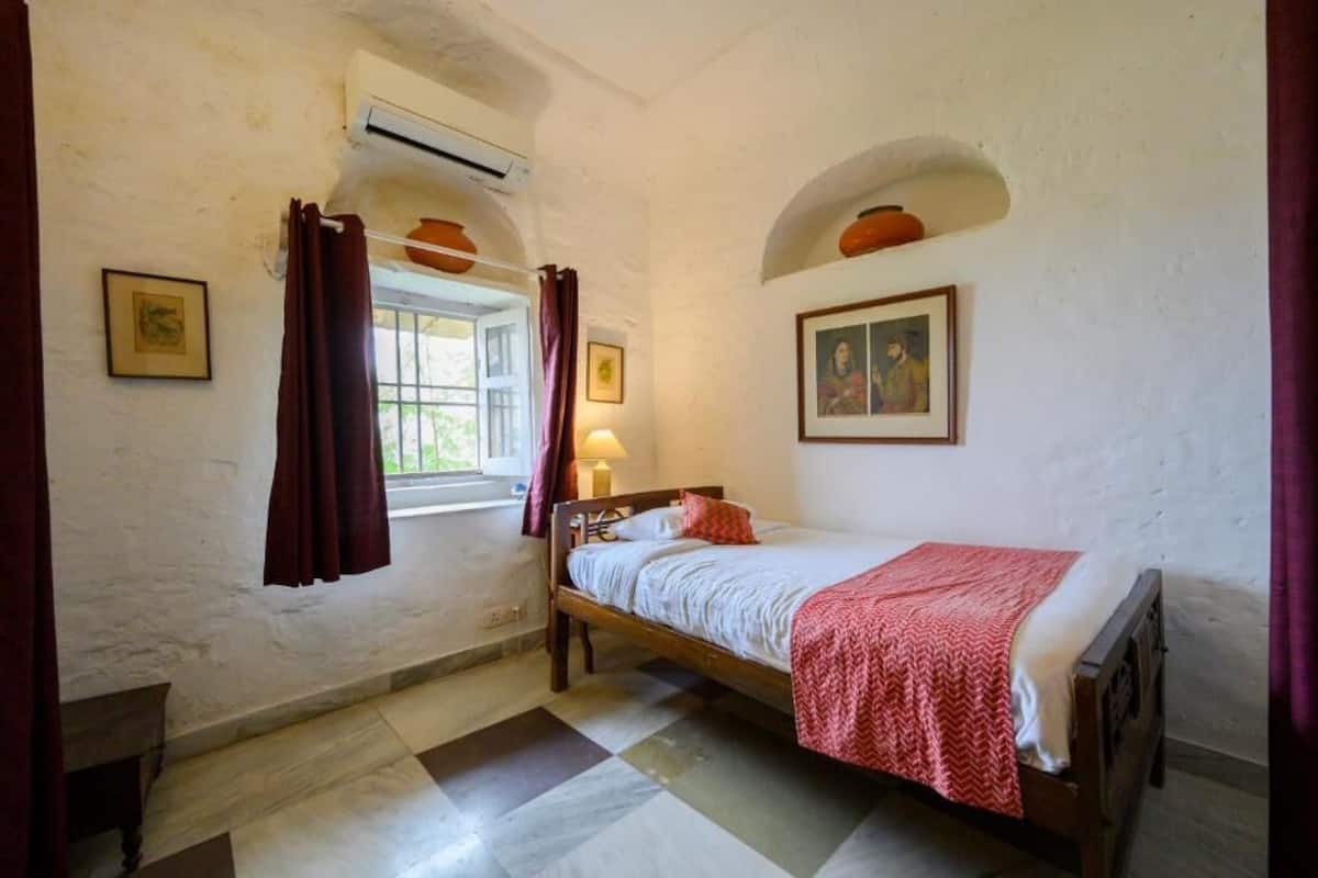 A bedroom with a bed, a window with curtains, a lamp, and a wall-mounted shelf.