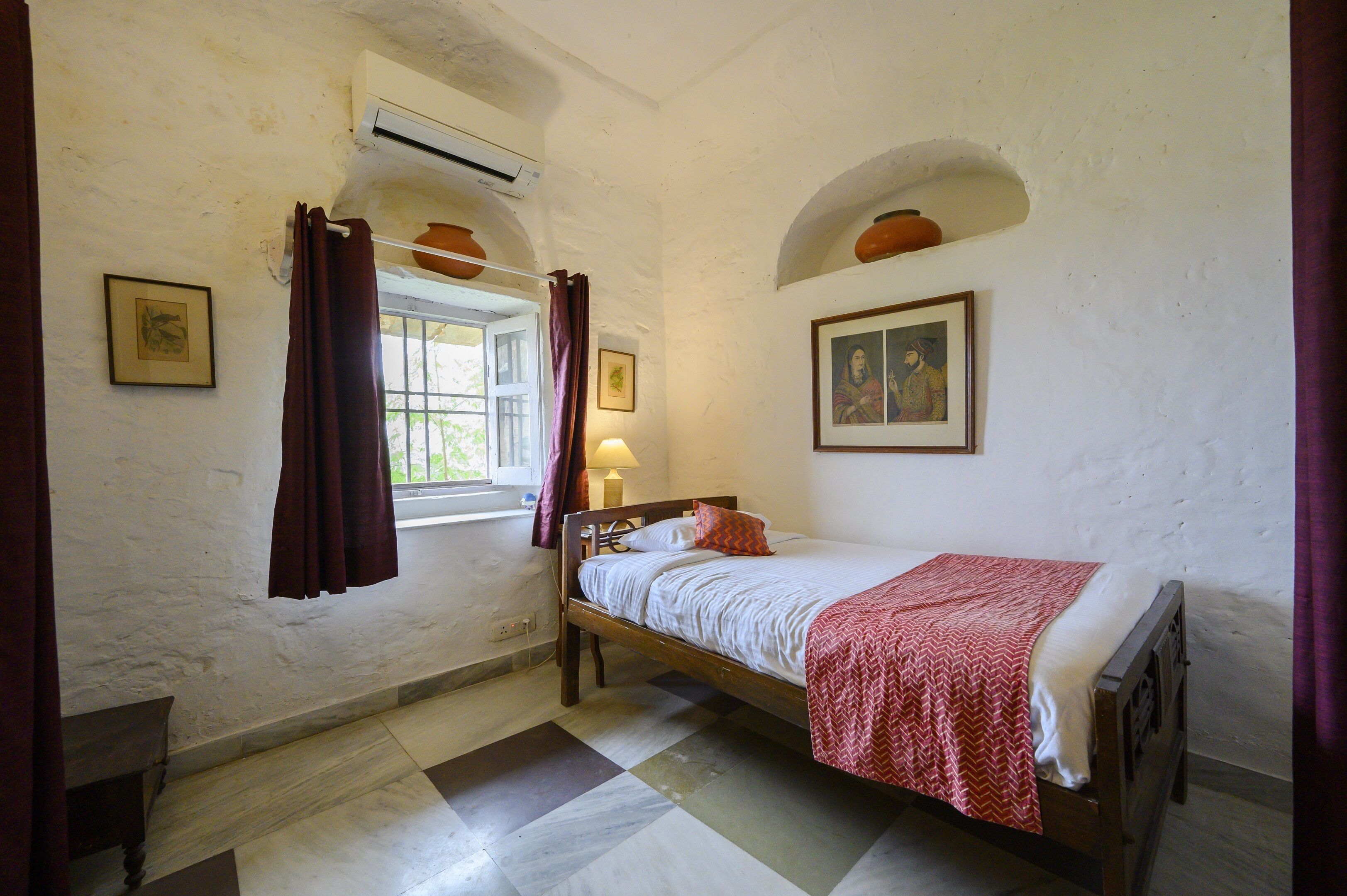 A room with a bed, a window with curtains, a wall-mounted shelf with pottery, and a framed picture.