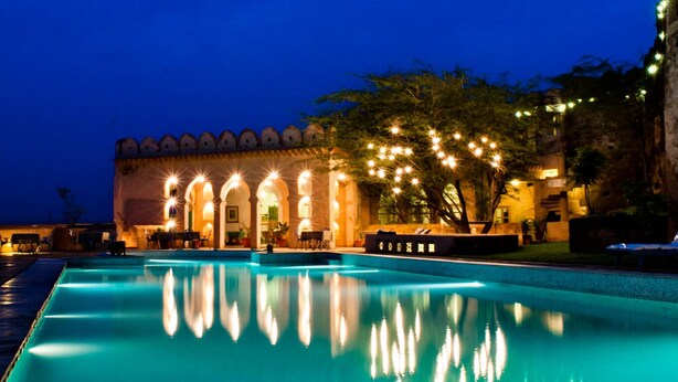 A pool area with a reflection of lights, a building with arches, and string lights. - Neemrana's - Hill Fort - Kesroli (Ramgarh)