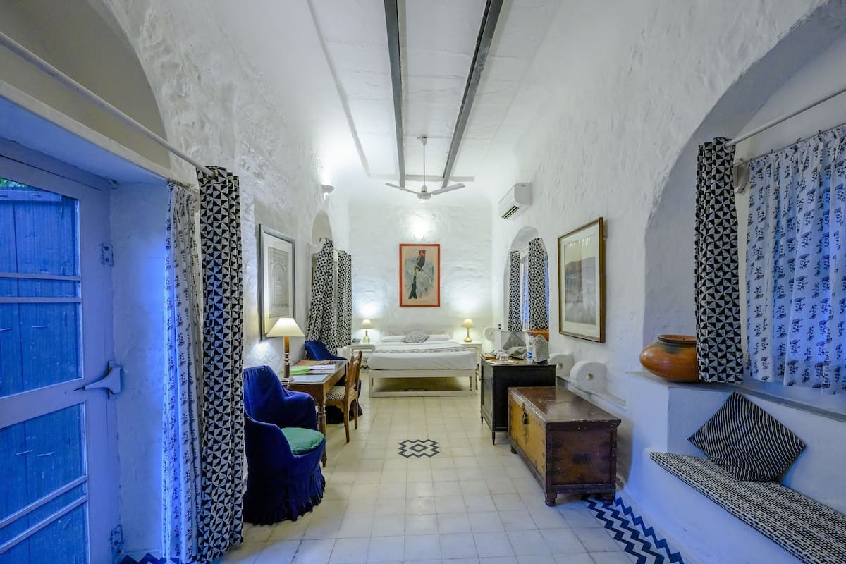 A narrow hallway leading to a bed with a headboard, a desk with a chair, a small table, and a bench. There are patterned curtains and a framed picture on the walls.