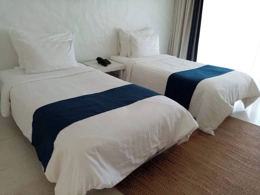 Two beds with white and blue bedding, a small bedside table with a phone, and a window with curtains.