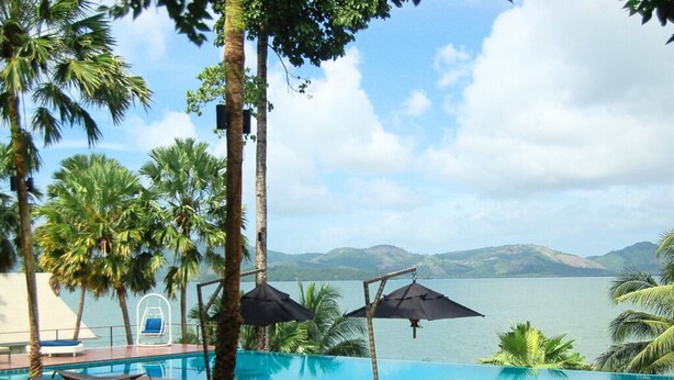 Zona junto a la piscina con sillones y sombrillas, con vistas a un cuerpo de agua y colinas.