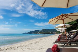 A beach with sun loungers and umbrellas, a clear sky, and a view of the sea and distant hills. - Anavana Beach Resort (Koh Samui)