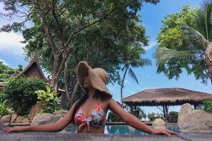 A woman in a swimsuit and hat by a pool with tropical trees and a thatched-roof structure in the background. - Anavana Beach Resort (Koh Samui)