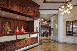 A reception area with a wooden reception desk, two staff members, a decorative plant, and a chandelier. - Anavana Beach Resort (Koh Samui)