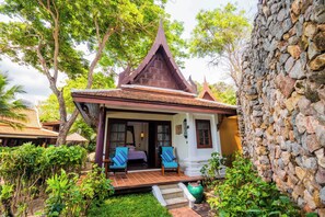 A traditional wooden house with a steeply pitched roof, a stone wall, and a porch with two chairs. - Anavana Beach Resort (Koh Samui)