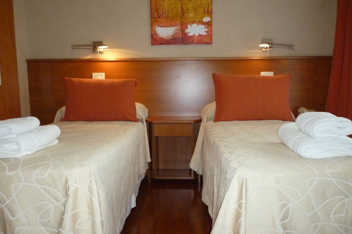 A hotel room with two beds, each with a white bedspread and orange pillows. There are two bedside tables with lamps, and a painting on the wall above the beds.