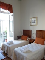 Habitación de hotel con dos camas, un balcón, un cabecero de madera, una mesita de noche con lámpara y un cuadro enmarcado en la pared.