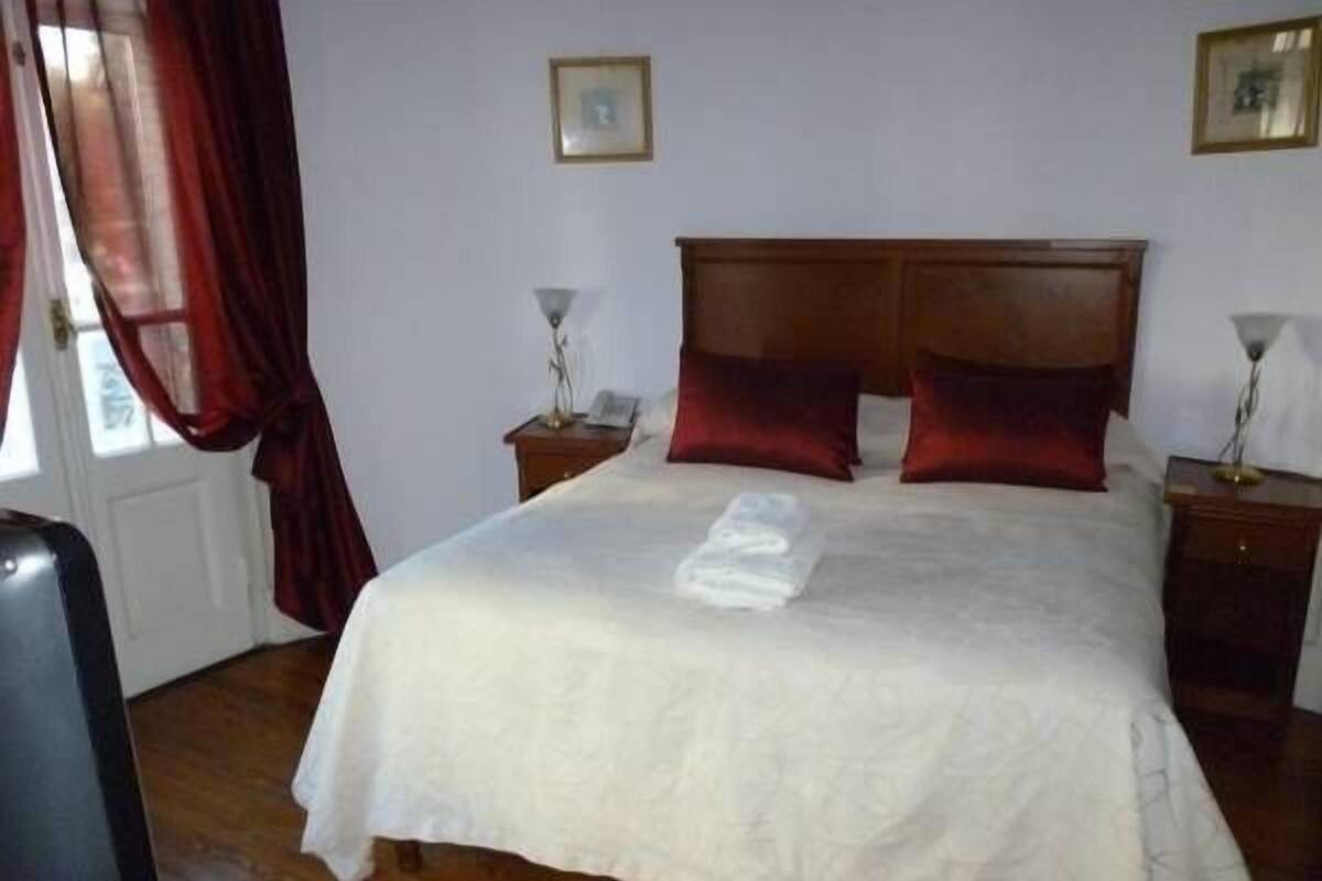 A neatly made bed with a white comforter and two red pillows, a wooden headboard, and a small bedside table with a lamp.