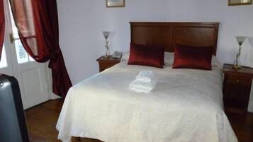 Una cama bien hecha con edredón blanco y dos almohadas rojas, cabecera de madera y una mesita de noche con una lámpara.