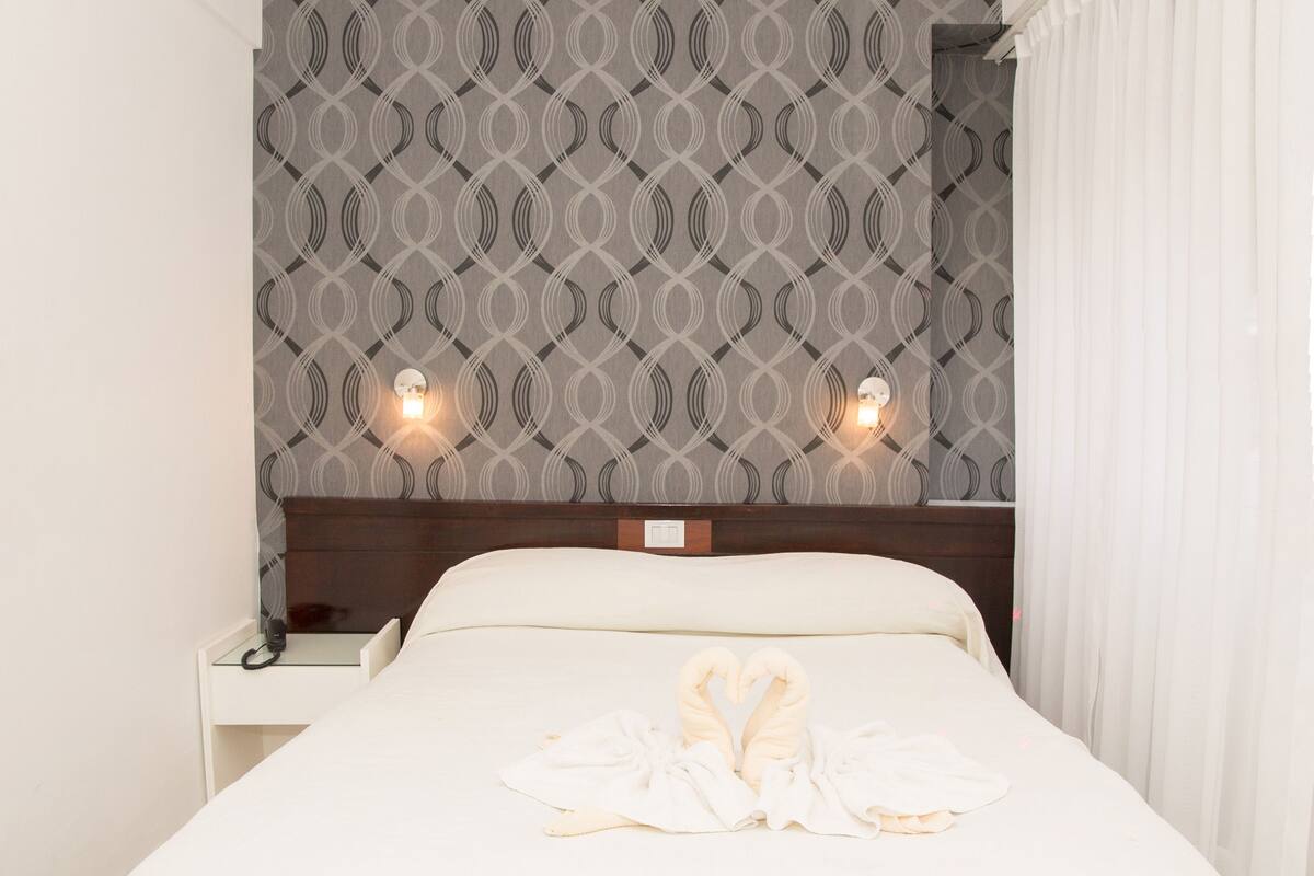 A neatly made bed with a heart-shaped towel arrangement, a nightstand with a lamp, and a patterned wallpaper.
