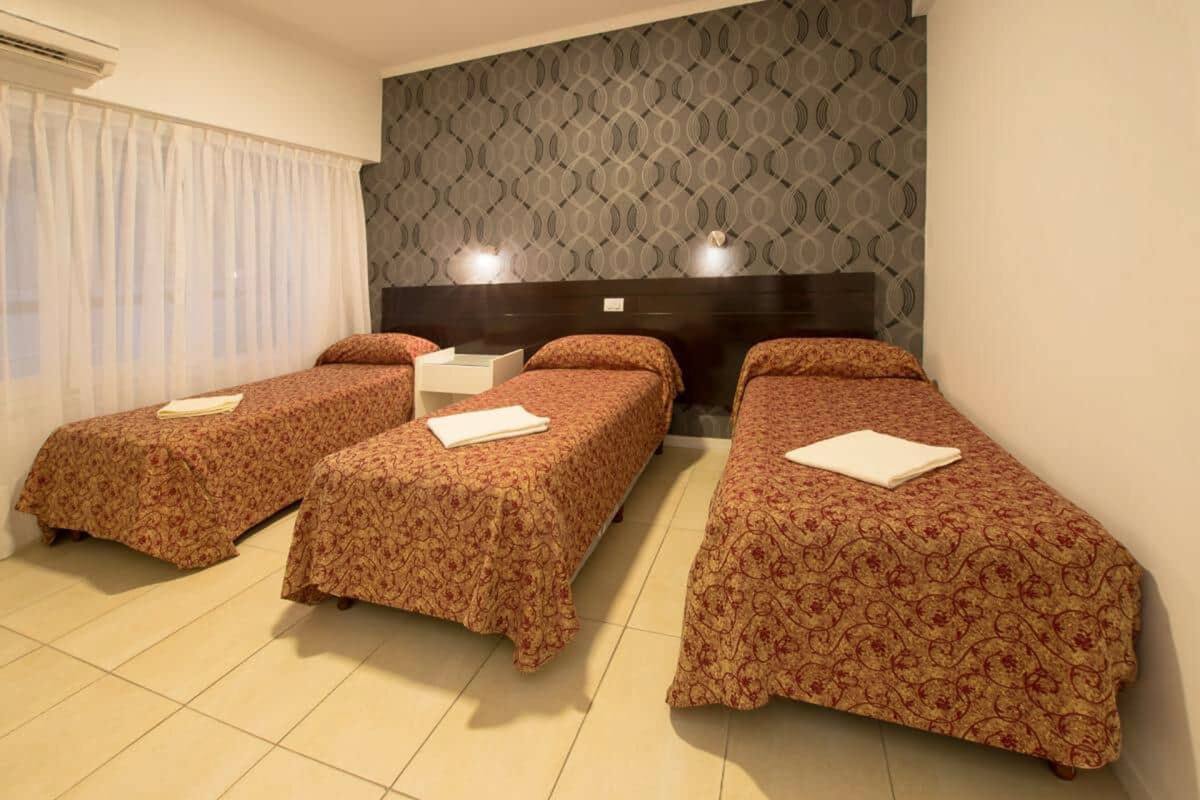 A hotel room with three single beds, a large window with curtains, a patterned wall, and a dark headboard.