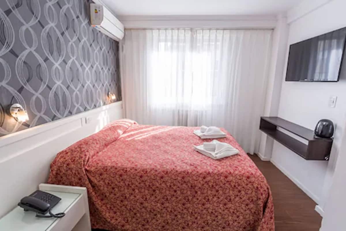 A hotel room with a bed, a bedside table with a phone, a TV mounted on the wall, and a window with curtains.