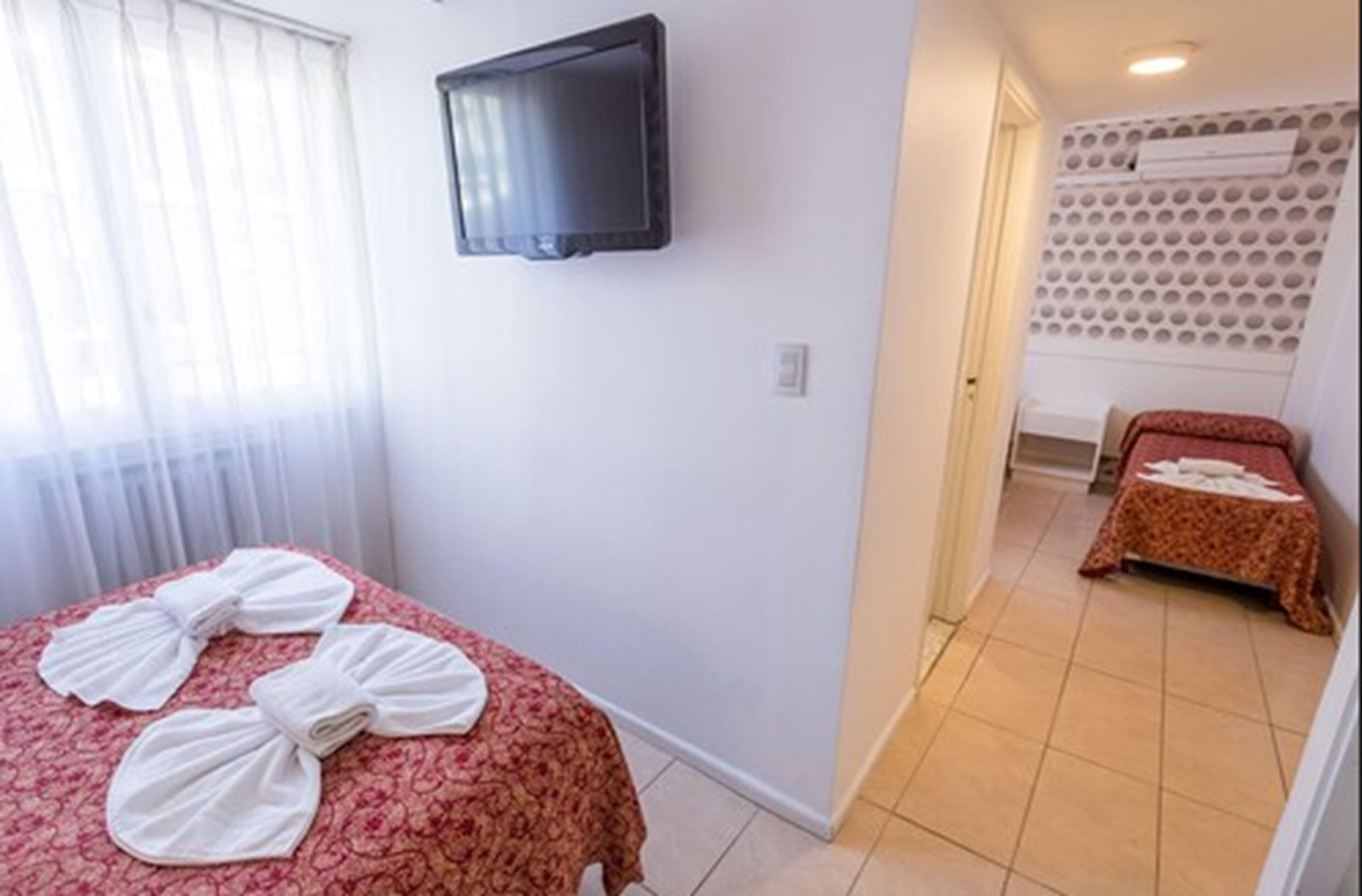 A hotel room with a bed, a TV mounted on the wall, a small table with a red tablecloth, and a separate bed with a red and white patterned bedspread.