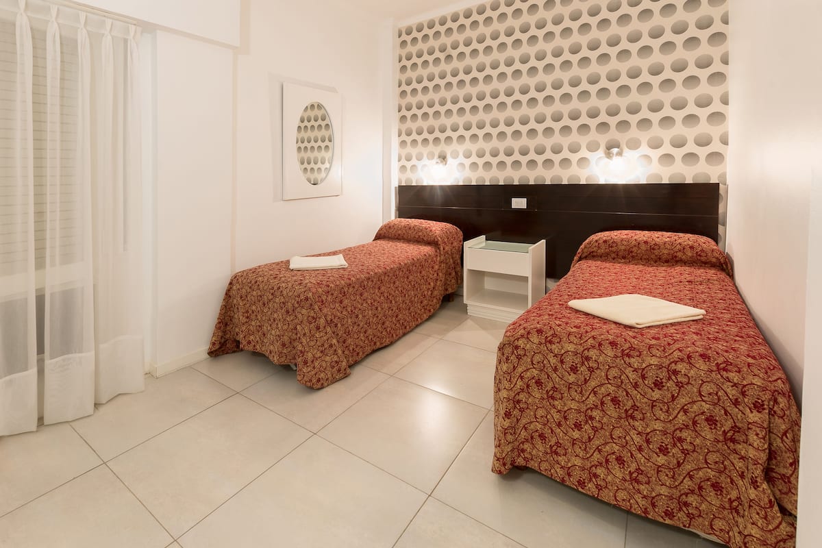 A hotel room with two beds, a large window with curtains, a wall-mounted light fixture, and a decorative wall panel.