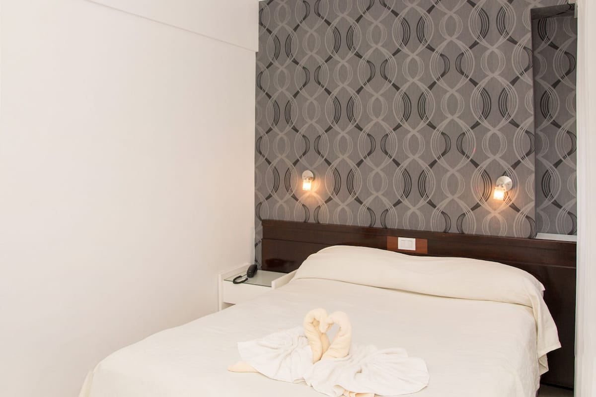 A neatly made bed with a white bedspread and two towels, a nightstand with a phone, and a patterned wallpaper on the wall.