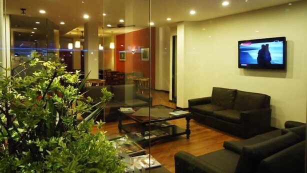 Un hall d’hôtel moderne doté d’un espace salon, d’un téléviseur fixé au mur et d’une cloison en verre qui sépare le hall de la salle à manger.