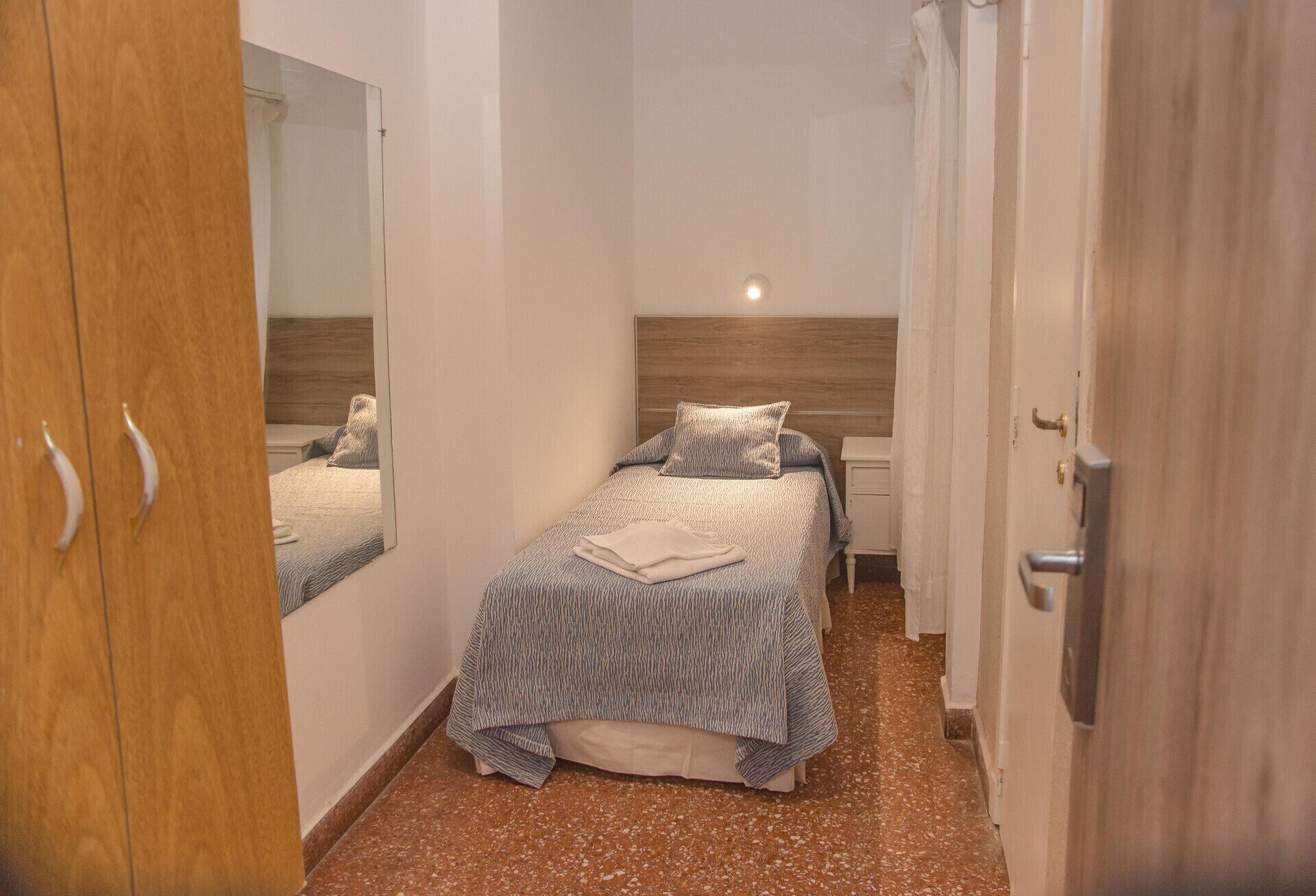 Quarto de hotel pequeno, com uma cama, um espelho, um guarda-roupa e uma porta.