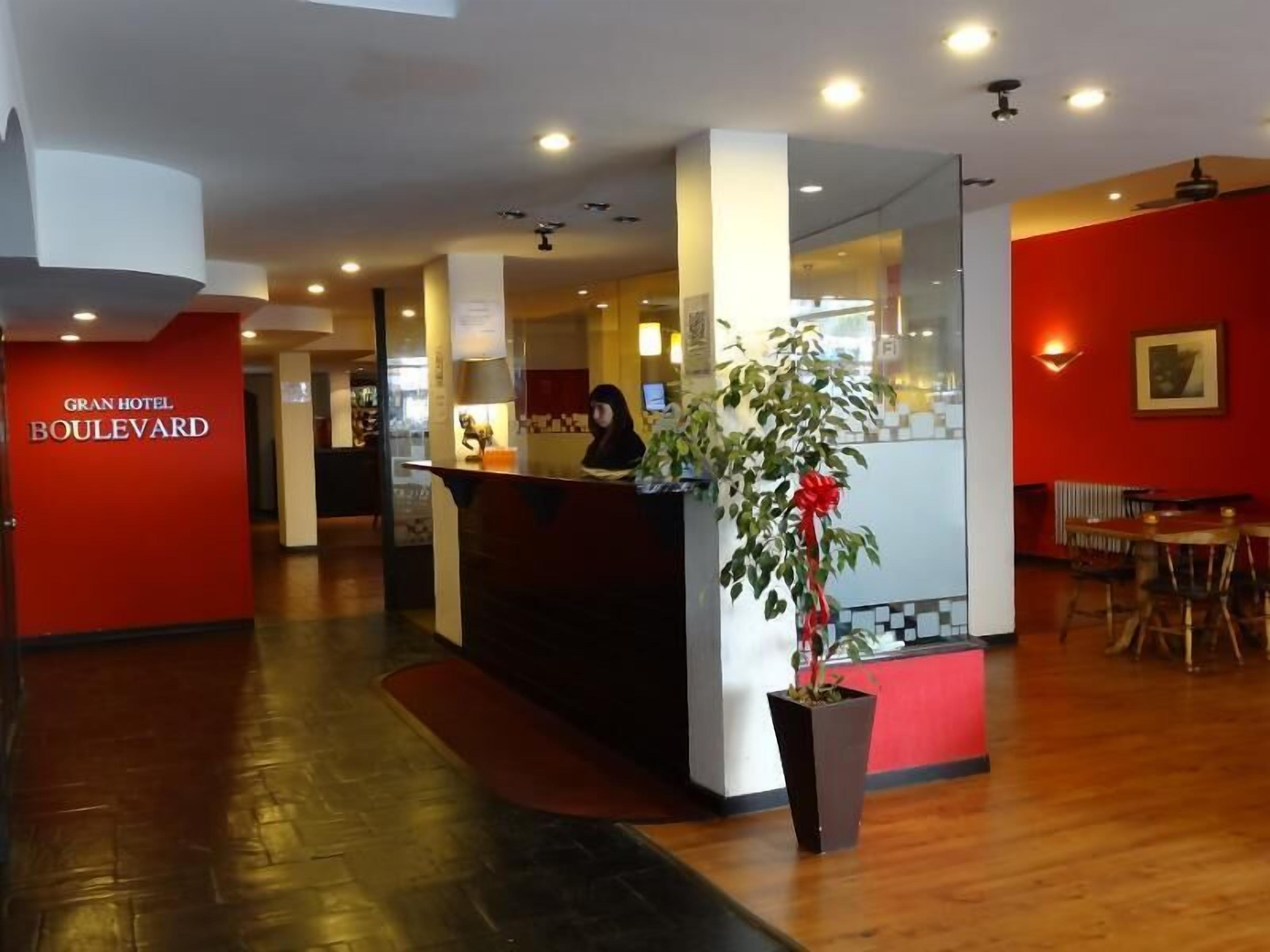 Zona de recepción de un hotel con pared roja, mostrador de recepción, recepcionista y planta en maceta con un lazo rojo.